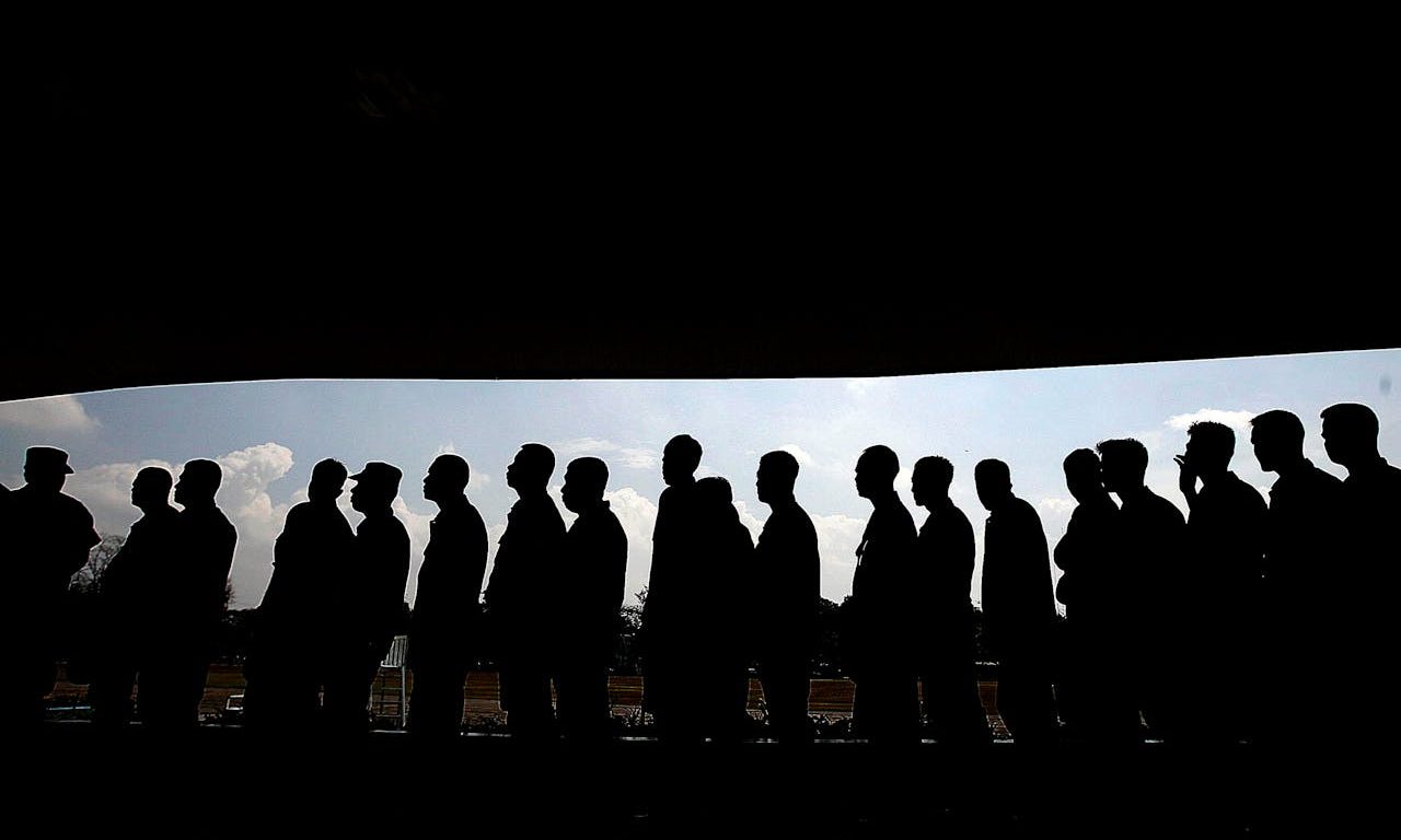 Una fila de personas en la ciudad. Se ven sus siluetas, dado que están iluminadas desde el fondo.