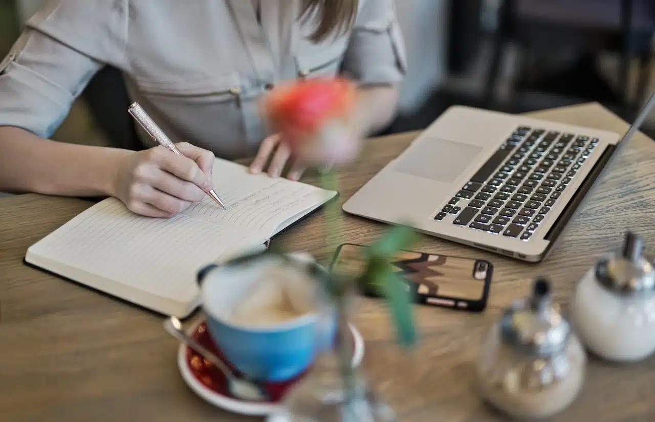 Mujer escribiendo en un cuaderno, junto a su ordenador portátil, una taza de café y una planta