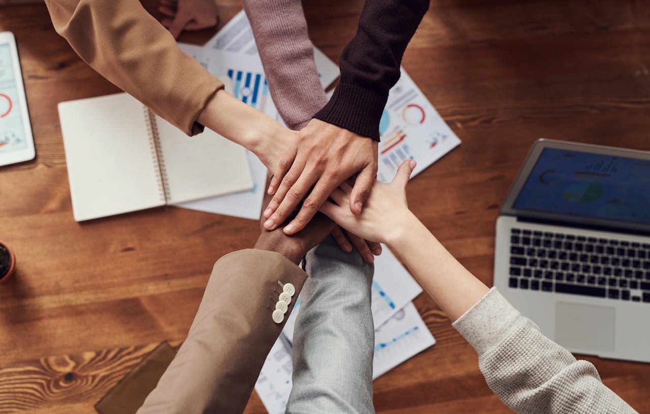 Personas diversas uniendo sus manos sobre una mesa de trabajo donde hay gráficos y un ordenador portátil