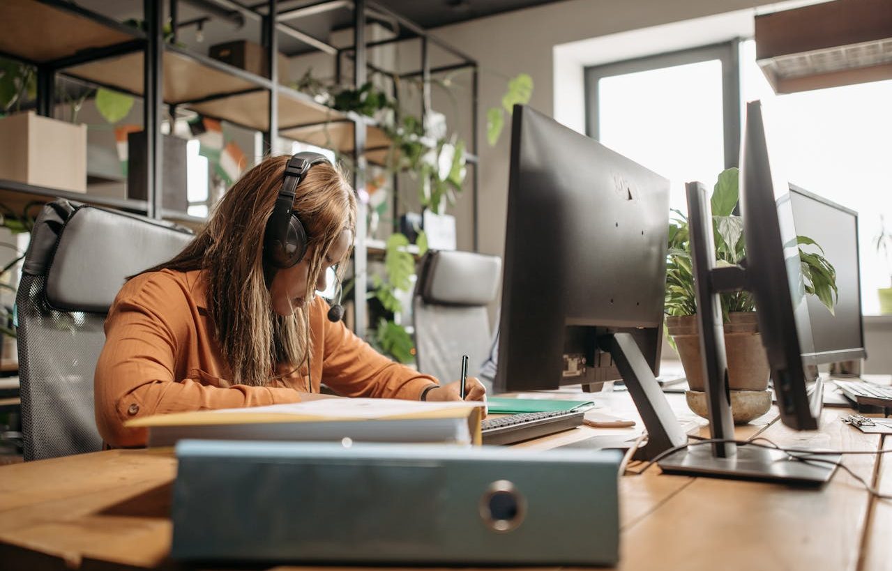 Mujer con auriculares tomando notas en un cuaderno frente al ordenador.